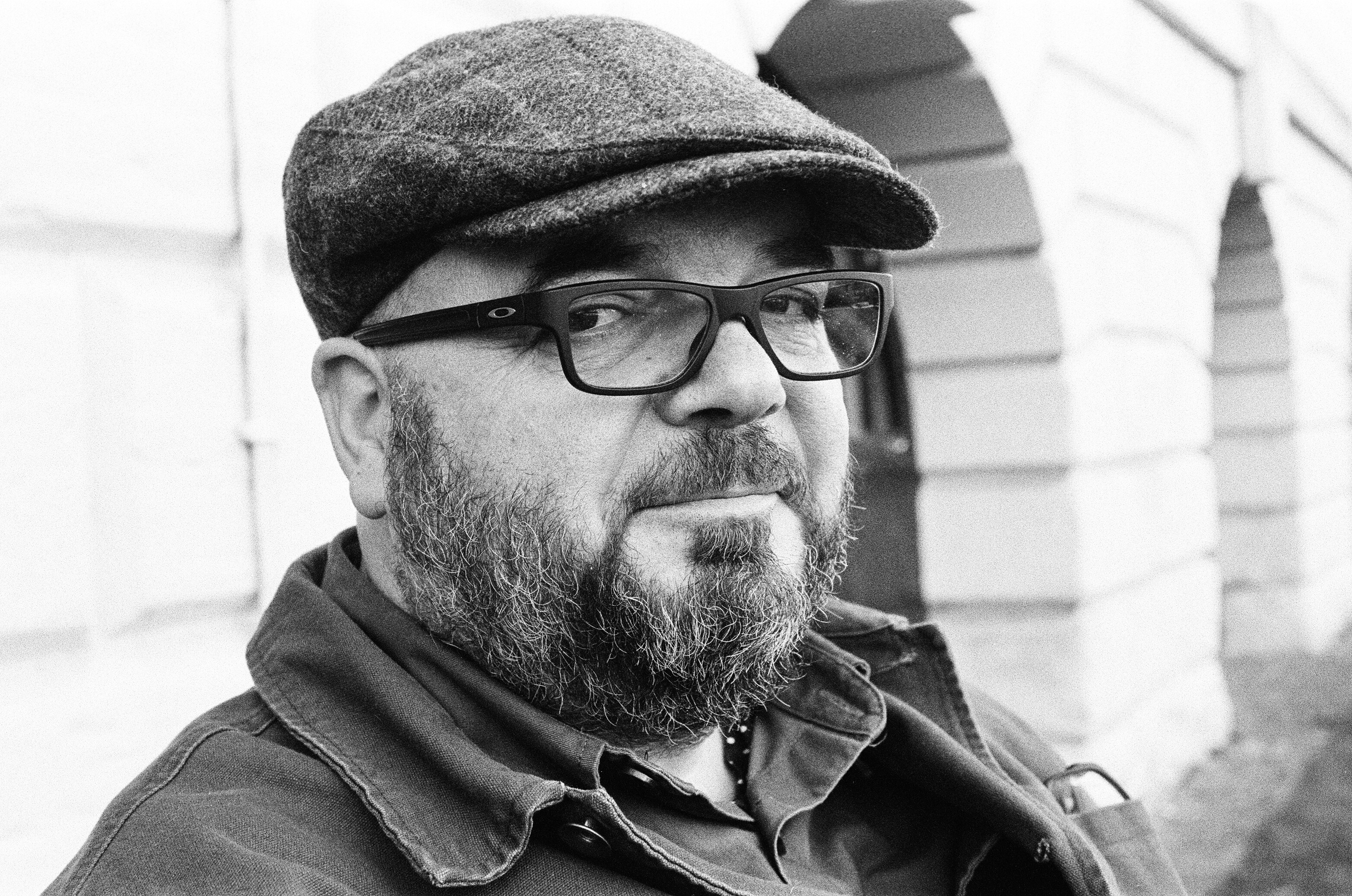 A black and white photo of Steve, a white middle-aged male, sitting on a bench in front of some stone arches. He has a flat cap on, glasses and a full beard.